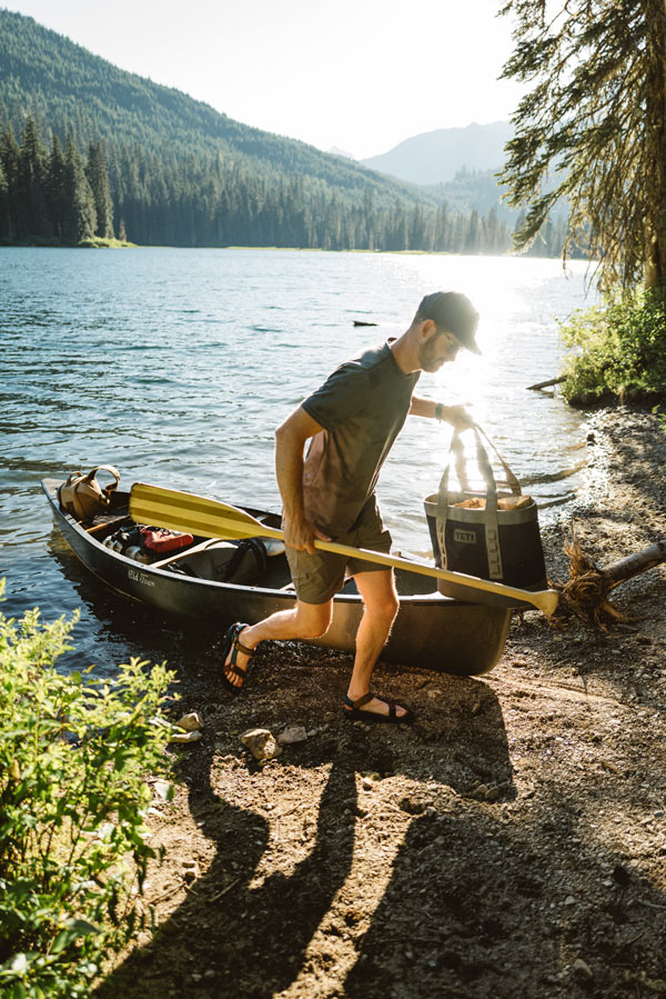 Ben walks up the shore of a lake from his canoe, carrying a bag of firewood. He wears Teva Hurricane XLT2 sandals.