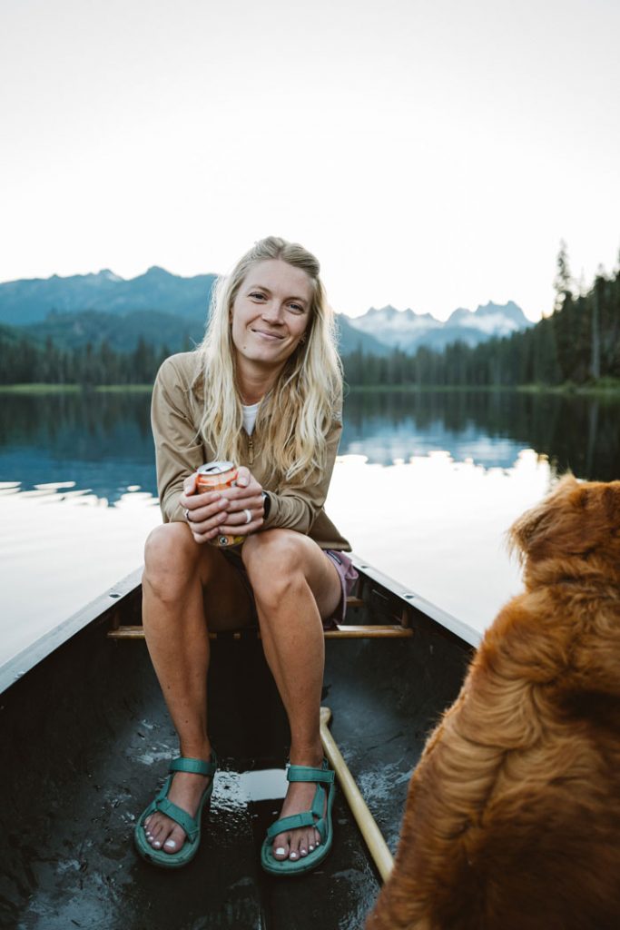 Whitney smiles in a canoe floating in a glassy lake, with her dog. She wears Hurricane XLT2 sandals.