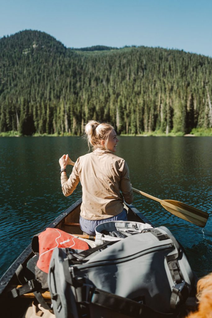 A view of Whitney paddling in a canoe on a calm lake, with all their camping gear inside and a forest of trees ahead.